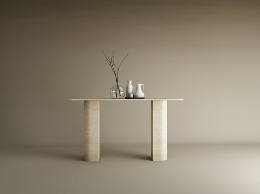 The TRAVERTABLE Rectangular Travertine Console Table with Sloped Pedestal Base in natural stone displays a glass vase with bare branches and two ceramic pitchers, set against a plain beige background.