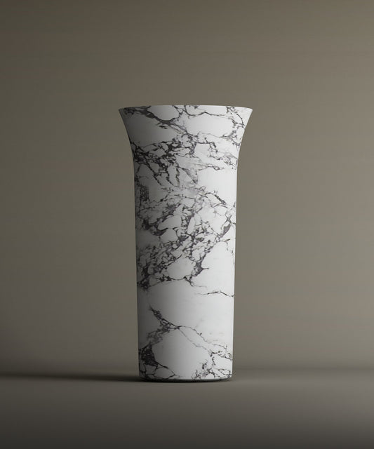 The TRAVERTABLE Calacatta Marble Cylinder Pedestal Vanity Sink features a tall, cylindrical white form with a flared rim and black-gray marble pattern, creating an elegant architectural statement against a neutral backdrop.