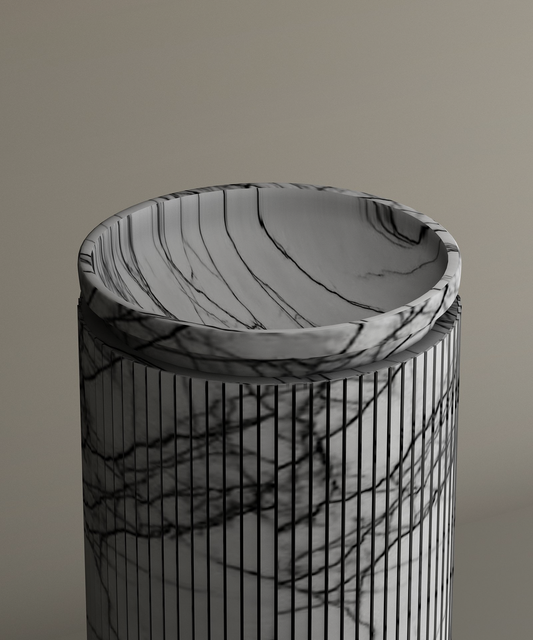 The TRAVERTABLE Lilac Round Marble Pedestal Vanity Sink features a cylindrical design with black veins and vertical lines, topped by a shallow circular dish. Set against a neutral beige background, it highlights sculptural elegance for your bathroom.